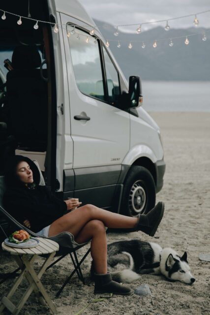 Ein Wohnmobil am Strand. Davor sitzt eine Frau im Campingstuhl im Pullover. Zu ihren Füßen liegt ein Hund.
